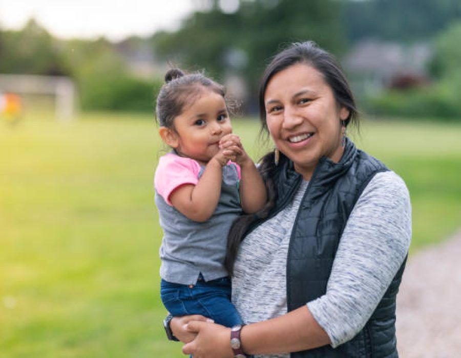 A Native American mom holds her toddler-age daughter affectionately while they take a break from walking in the park to smile at the camera.