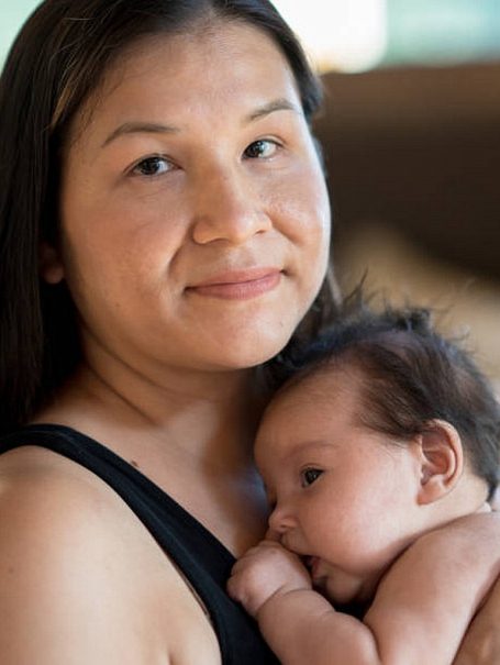 An indigenous mother sits with her newborn daughter on her chest as they enjoy some skin-on-skin time together.  The mother is dressed casually in a black tank top and the baby is wearing just a diaper as they share the intimate moment together.